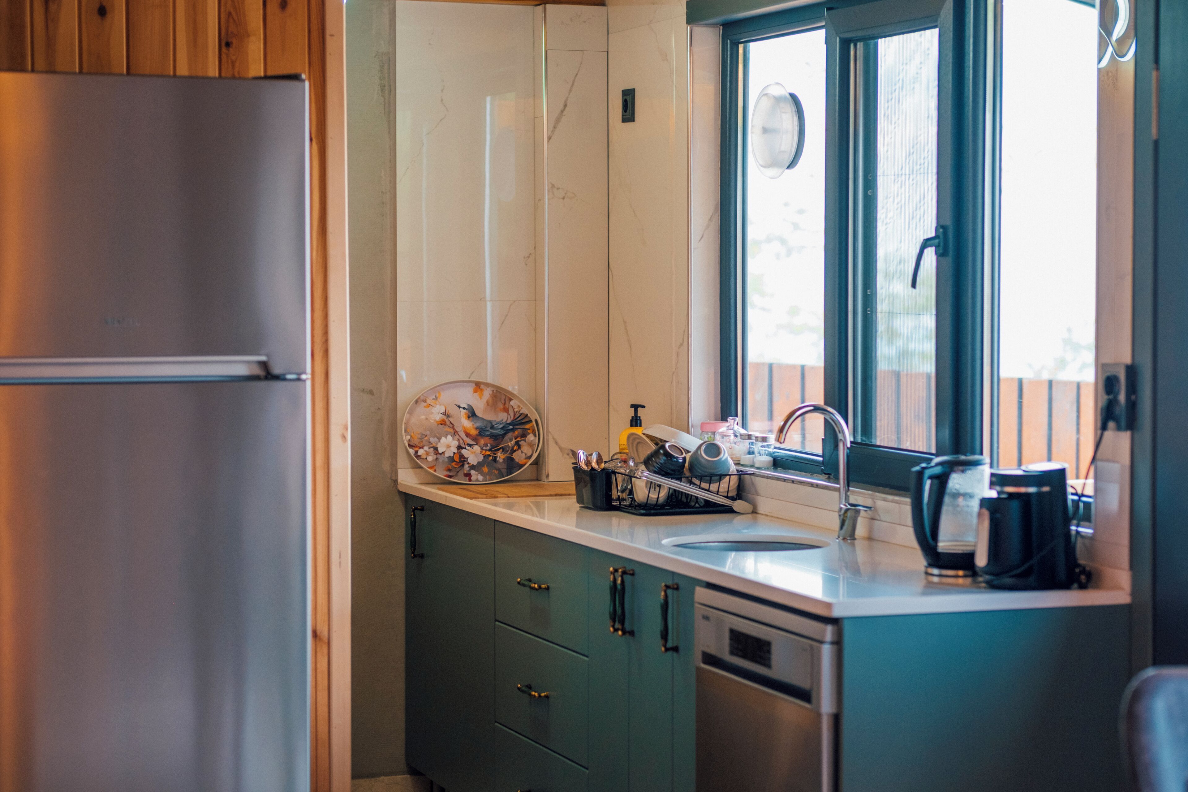 A kitchen with a stainless steel refrigerator, a window with a view, a sink with a faucet, and a coffee maker.