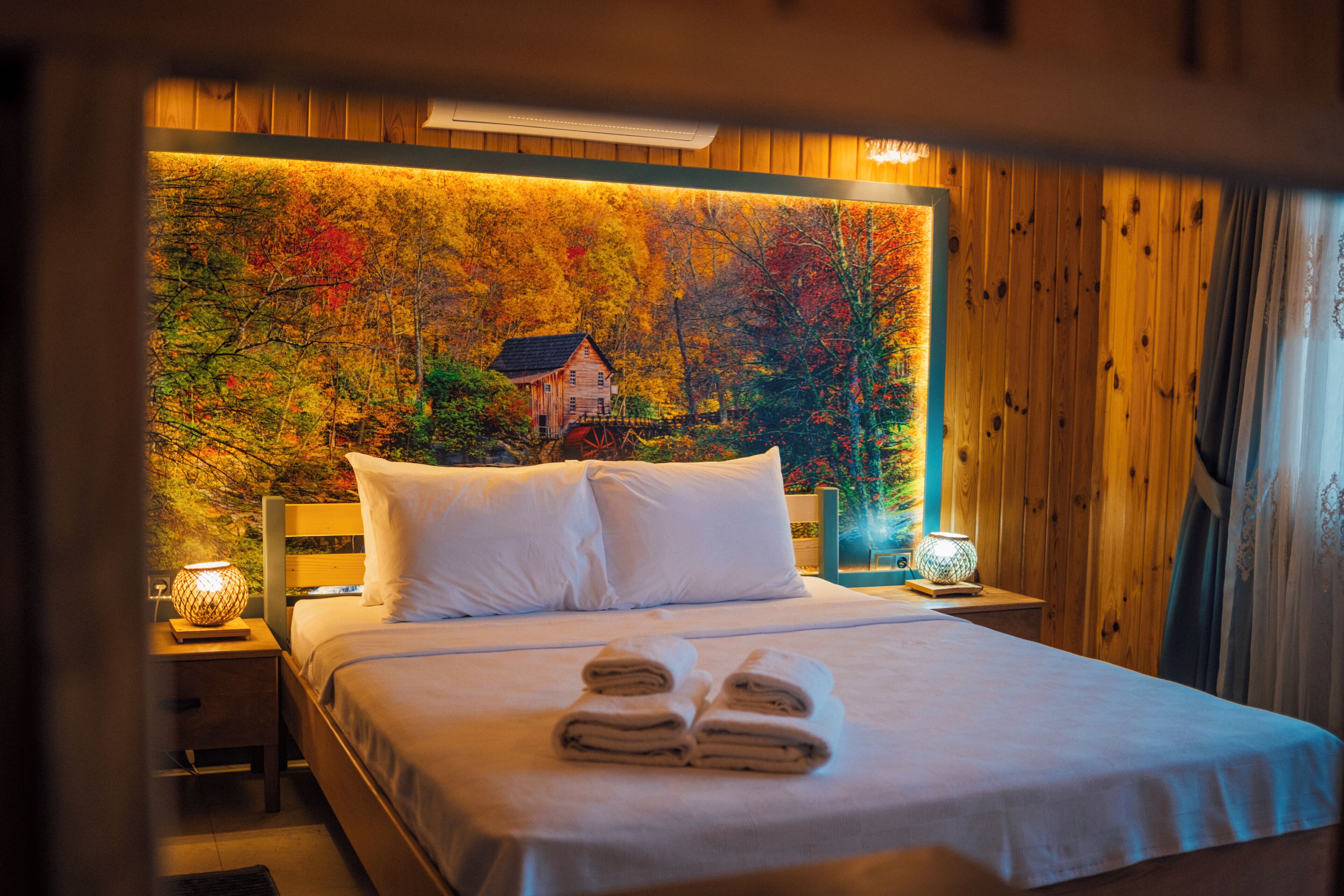 A neatly made bed with white linens and two rolled towels, a wooden headboard, and a nightstand with a lamp.