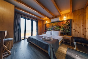 A wooden cabin-style room with a large bed, a bench, a desk, and a view through a large window.