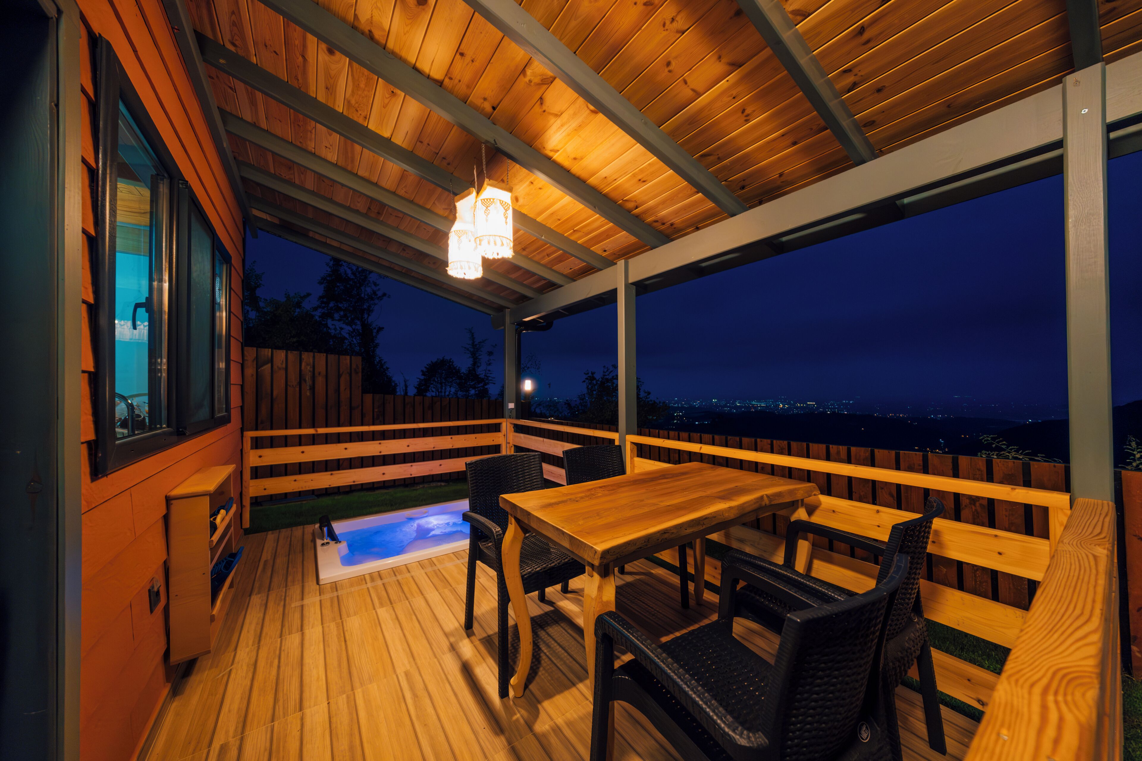 A wooden deck with a table and chairs, a hot tub, and a view of the city at night.