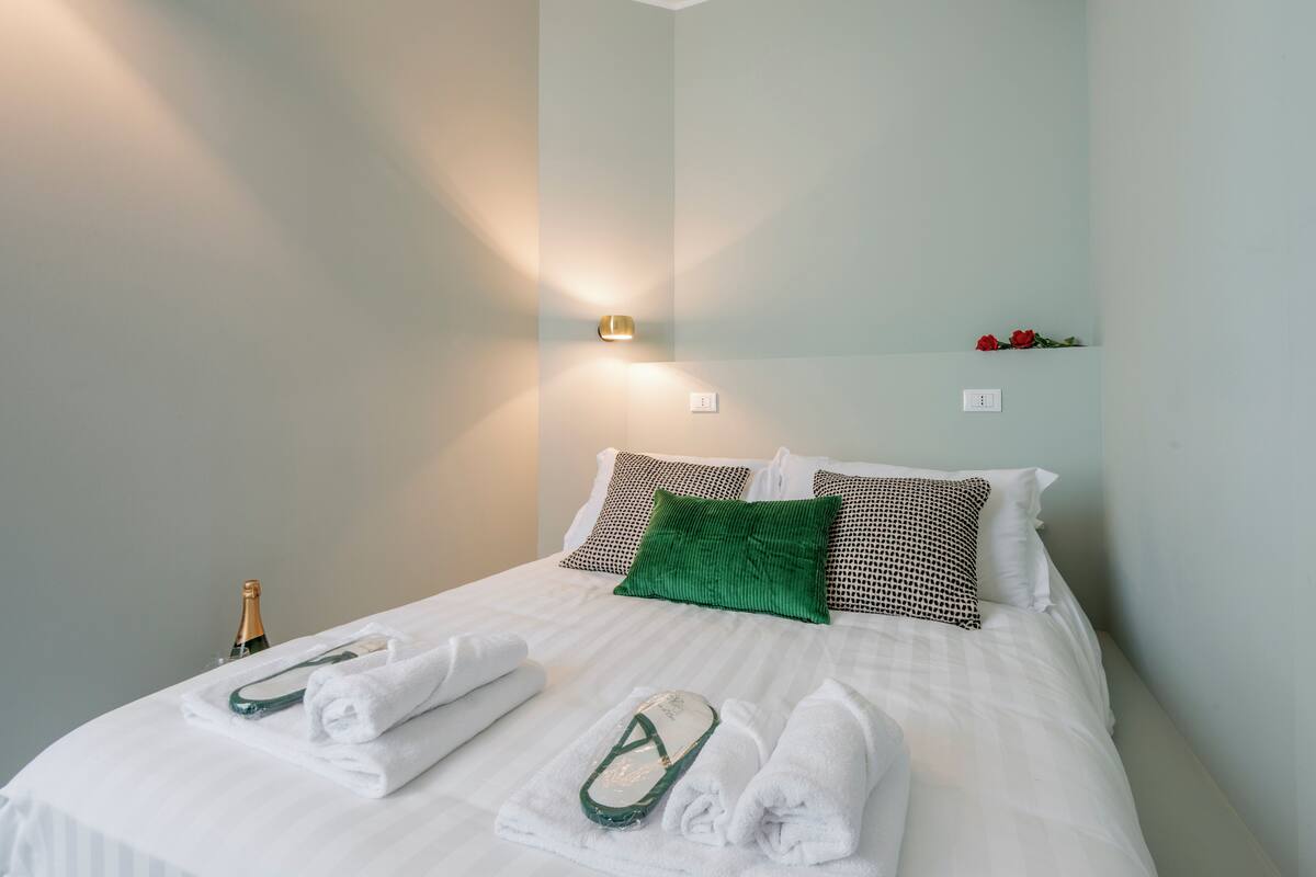 Un lit bien fait, avec des draps blancs, un oreiller vert et une bouteille de champagne posée à côté.