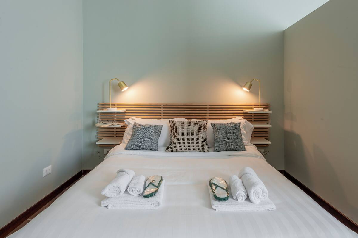 Un lit bien fait, avec deux jeux de serviettes et des sandales, une tête de lit en bois dotée d’un éclairage intégré et un mur de couleur claire.