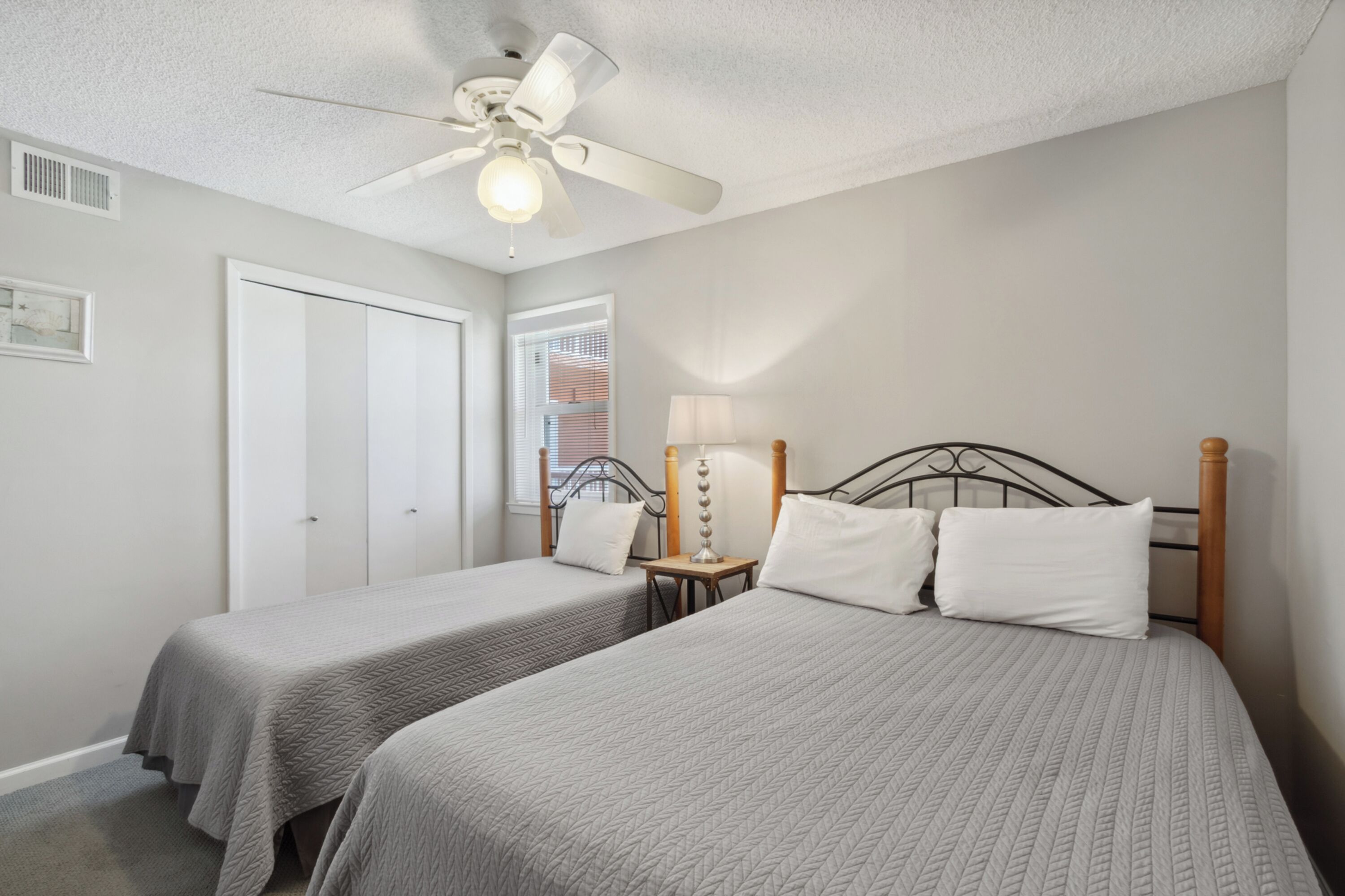 A bedroom with two beds, a ceiling fan, a nightstand with a lamp, and a window with blinds.