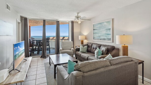 A living room with a sofa, armchairs, a coffee table, and a TV. There is a balcony with a view of the ocean.