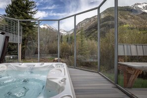 A hot tub with a mountain view, surrounded by glass walls and a wooden deck.