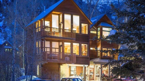 A two-story wooden cabin with large windows and a balcony, surrounded by trees and snow.