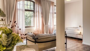 A modern living room with a sofa, a coffee table, and a lamp. There is a large window with curtains, and a plant is visible in the foreground. - Studio 9 (Fulda)