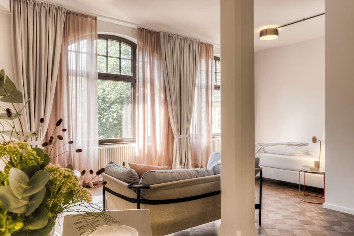 Un soggiorno moderno con un divano, un tavolino e una lampada. Si vede un'ampia finestra con tende, e una pianta sullo sfondo.