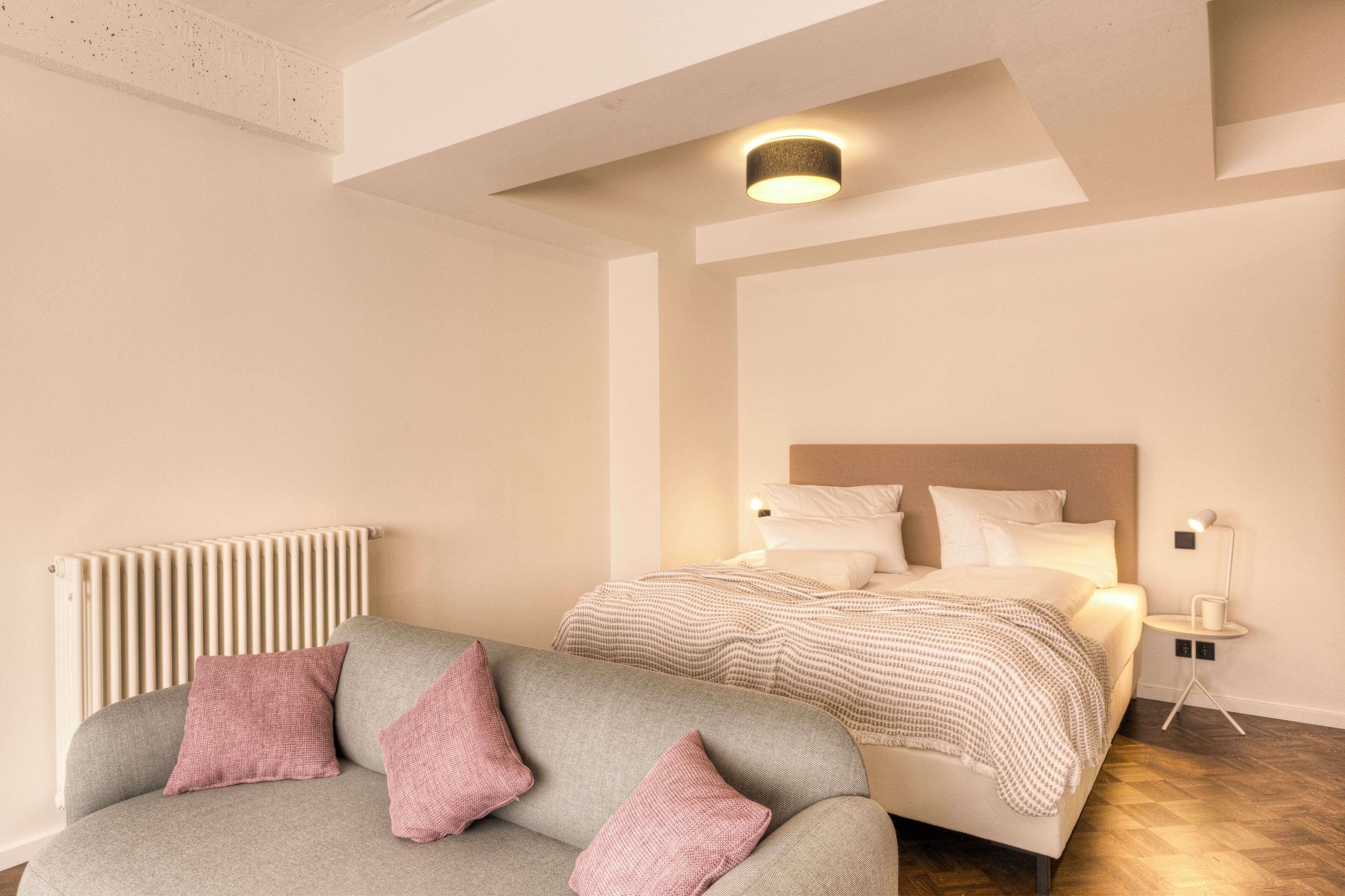 A modern bedroom with a bed, a grey sofa with pink cushions, a bedside table with a lamp, and a radiator.