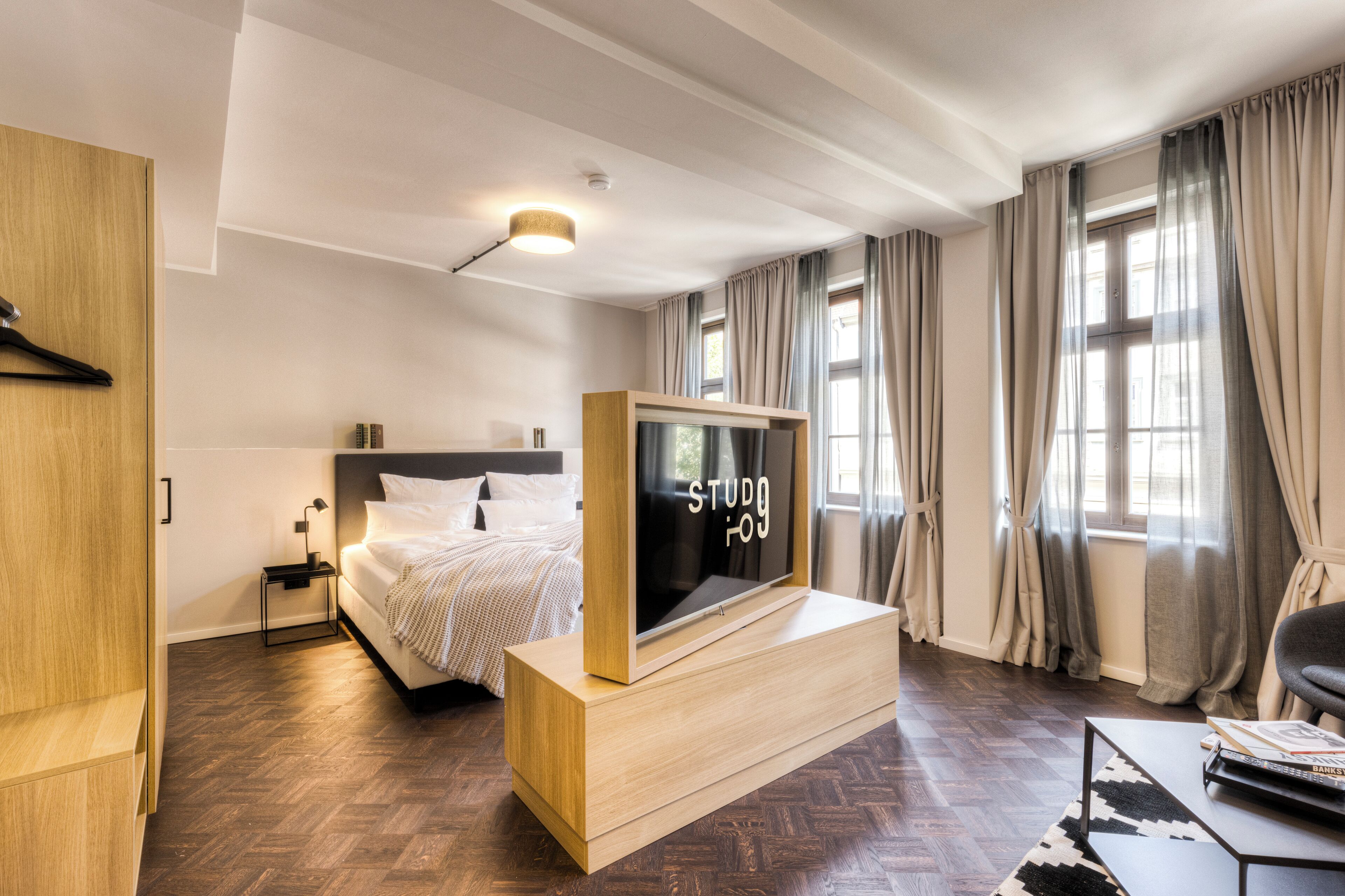 A modern hotel room with a large bed, a flat-screen TV, a wooden bedside table, and a view of the city through large windows.