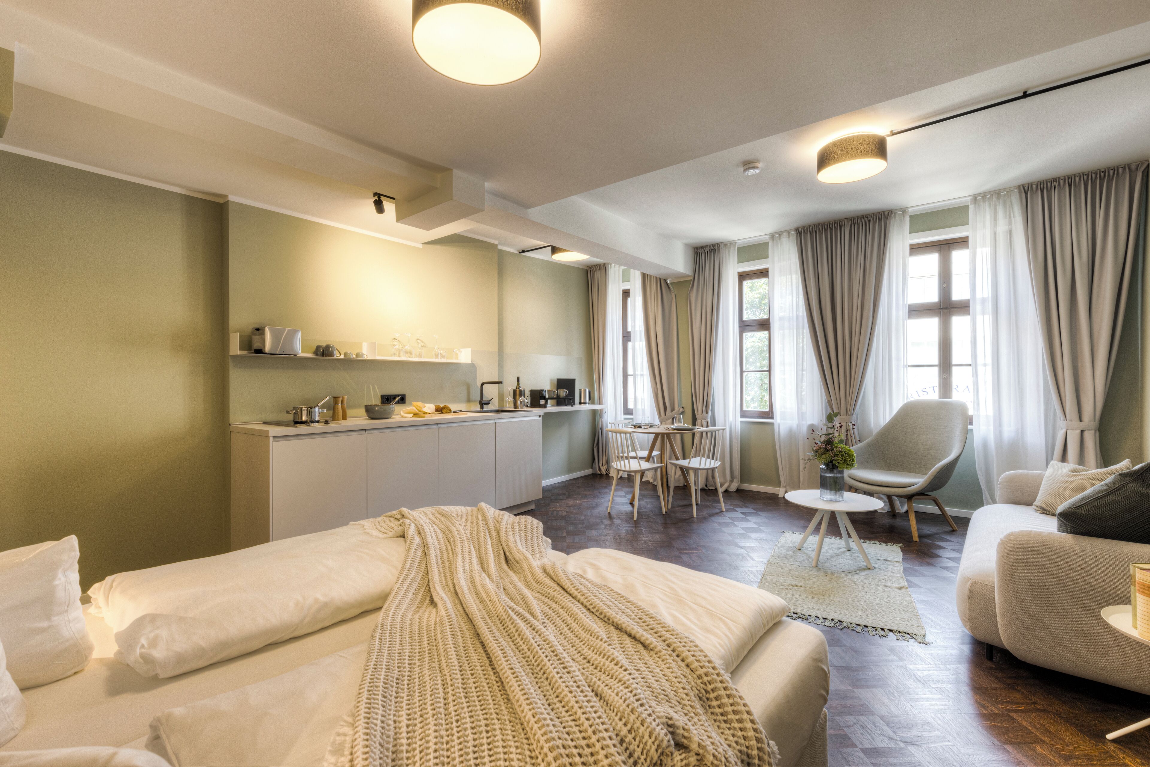 A modern hotel room with a bed, a sofa, a dining area, and a kitchen.