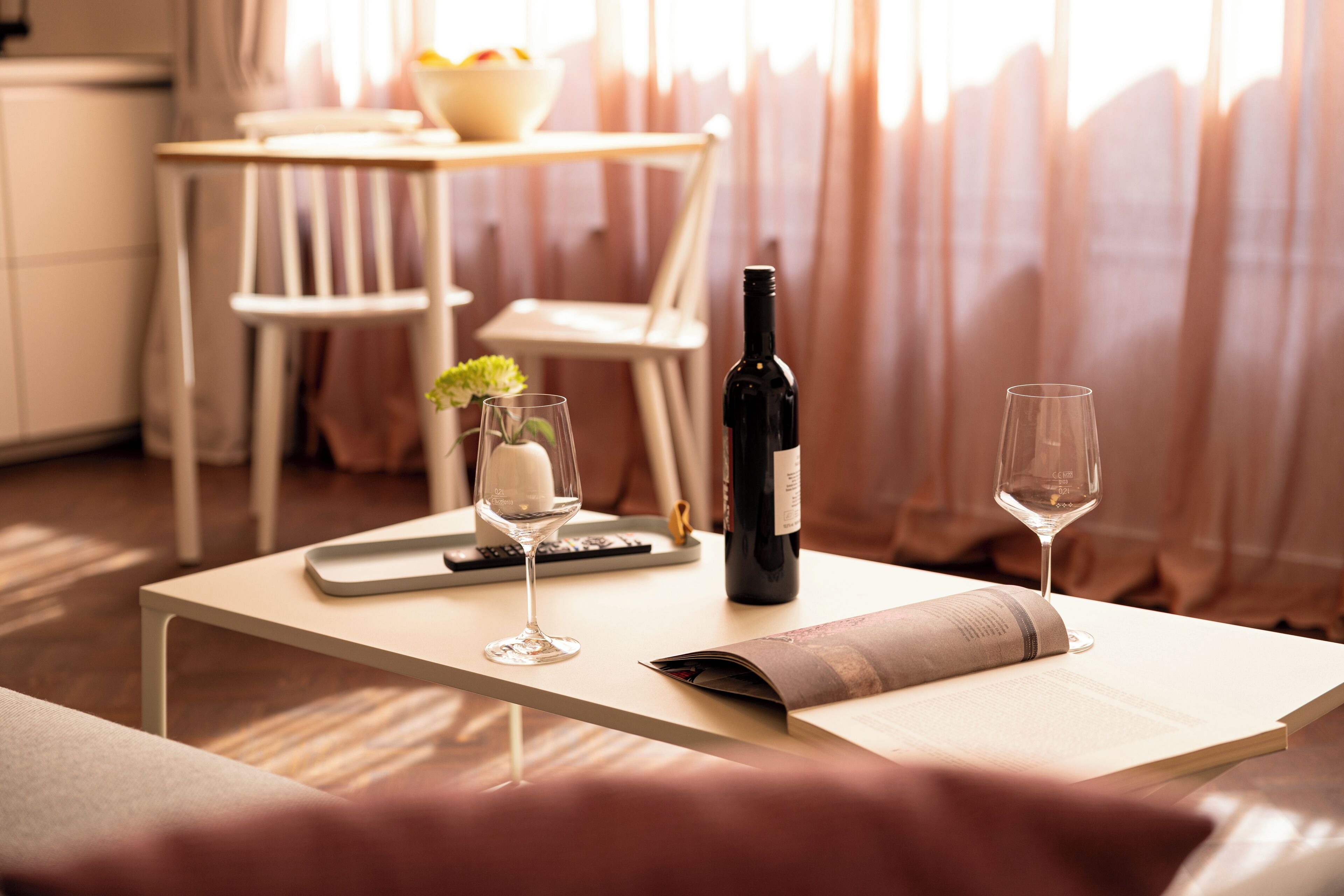 A dining table with a wine bottle, two wine glasses, a bowl of fruit, and a magazine.