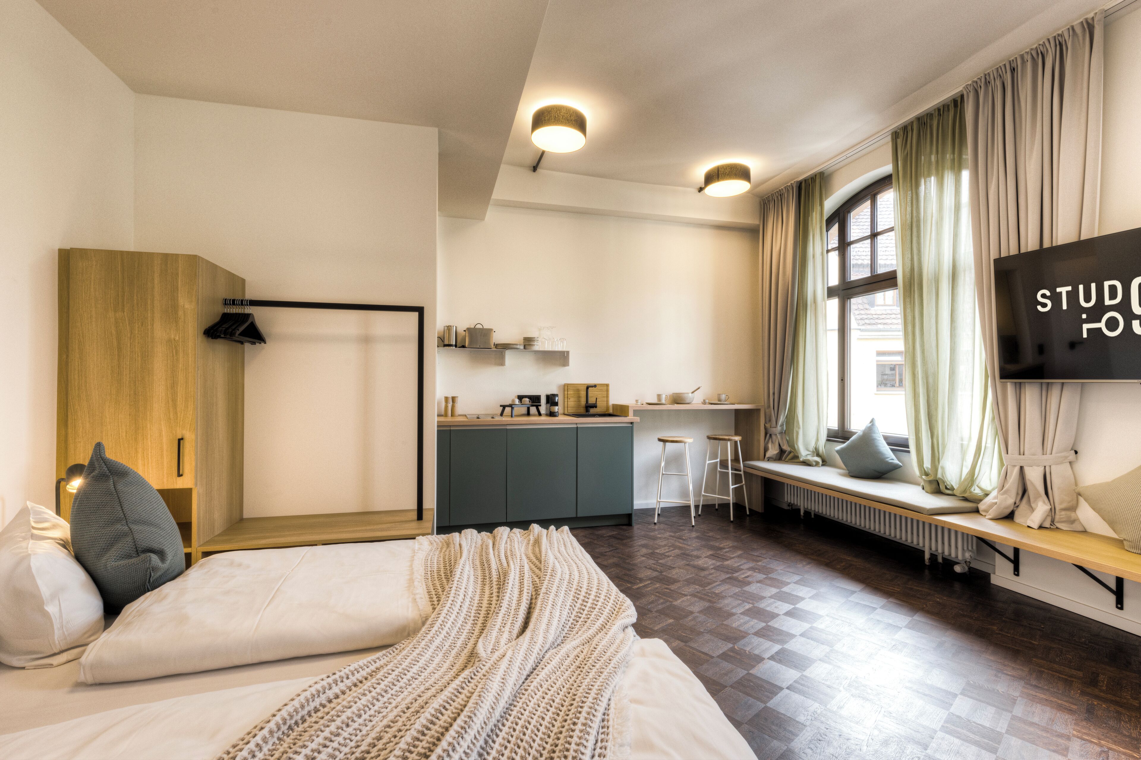 A modern hotel room with a bed, a wooden wardrobe, a kitchenette, and a large window with curtains.