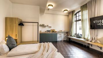 A modern hotel room with a bed, a wooden wardrobe, a kitchenette, and a large window with curtains.