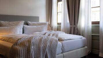Una cama bien hecha con sábanas blancas y una manta de punto, una mesita de noche y una ventana con cortinas.