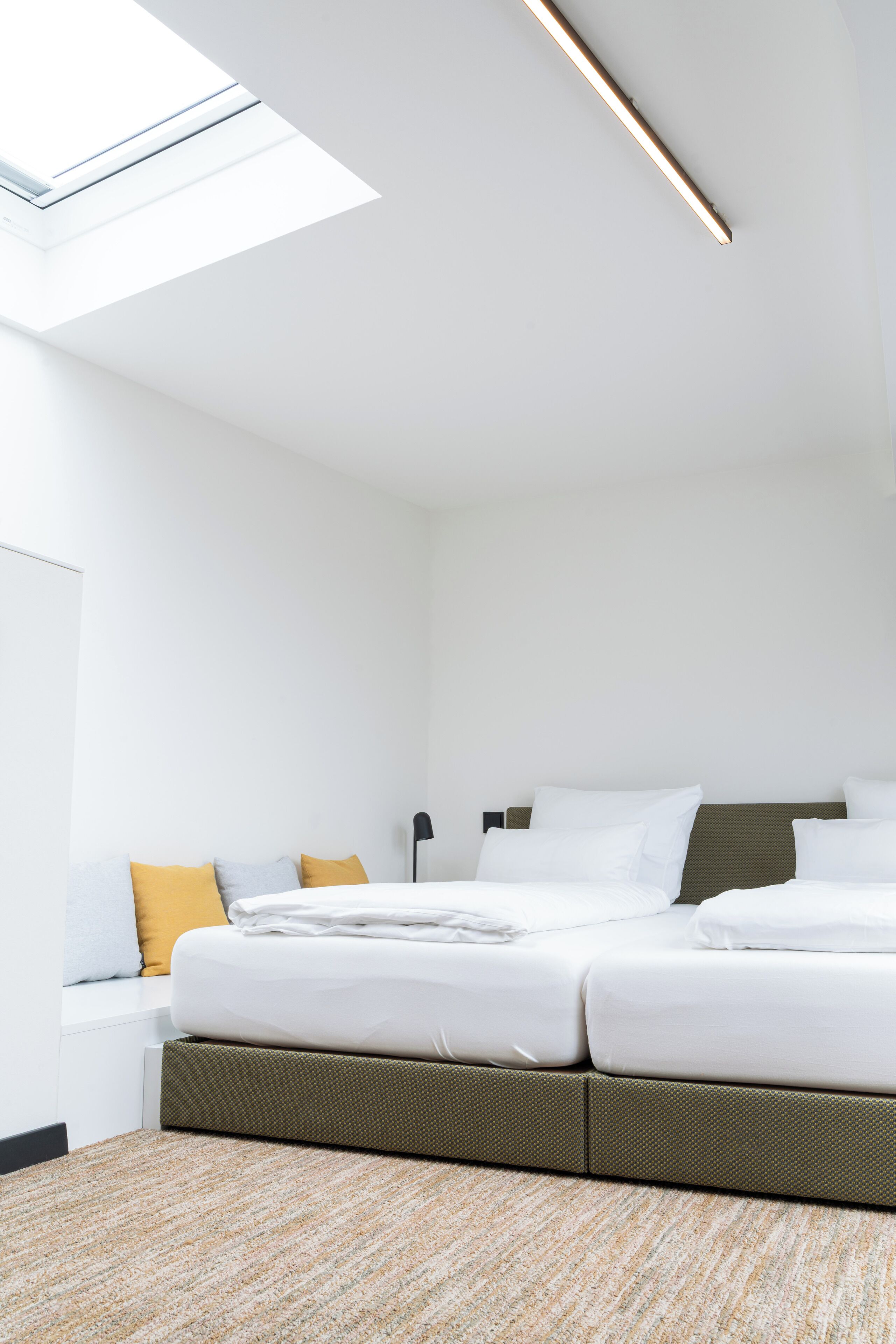 Quarto de hotel moderno com claraboia, cama com roupa de cama branca, cabeceira verde e carpete texturizado.