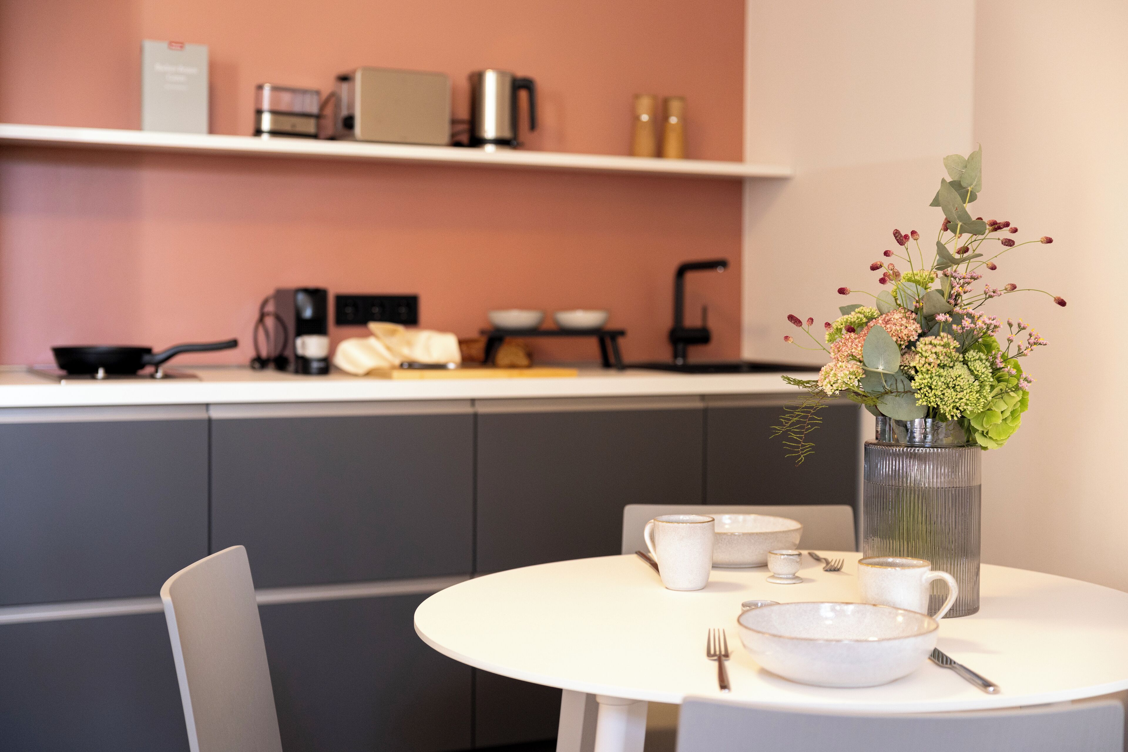 A modern kitchen with a white countertop, a coffee machine, and a vase with flowers on the table.