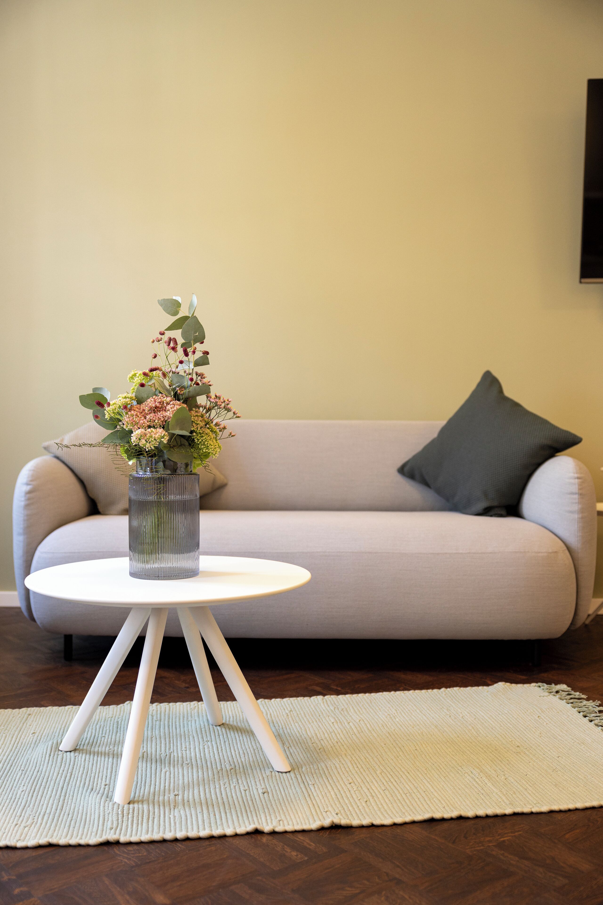 A living room with a grey sofa, a round white coffee table with a vase of flowers, and a textured rug on the floor.