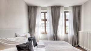 A bedroom with a large bed, white bedding, grey curtains, and a window with a view of buildings. - Studio 9 (Fulda)
