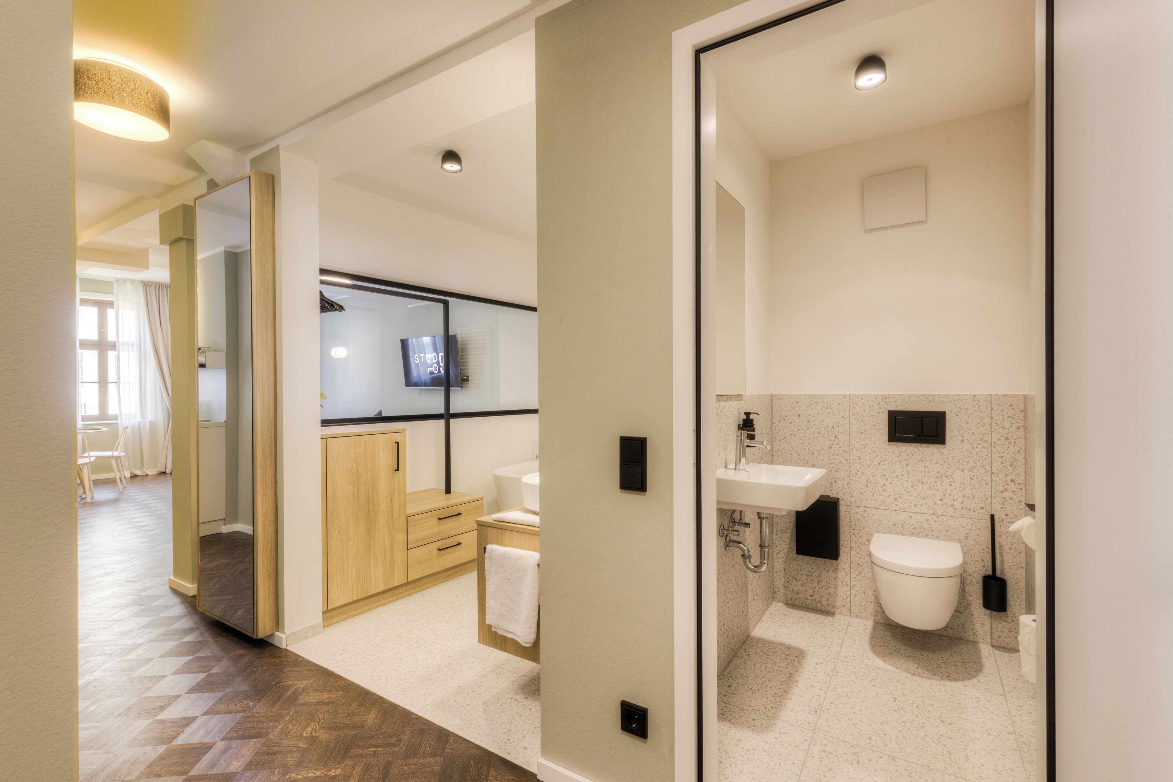Une salle de bain moderne avec des toilettes, un lavabo et un meuble-lavabo. On y trouve un meuble en bois et un miroir. Le sol est recouvert d’un motif en chevrons.