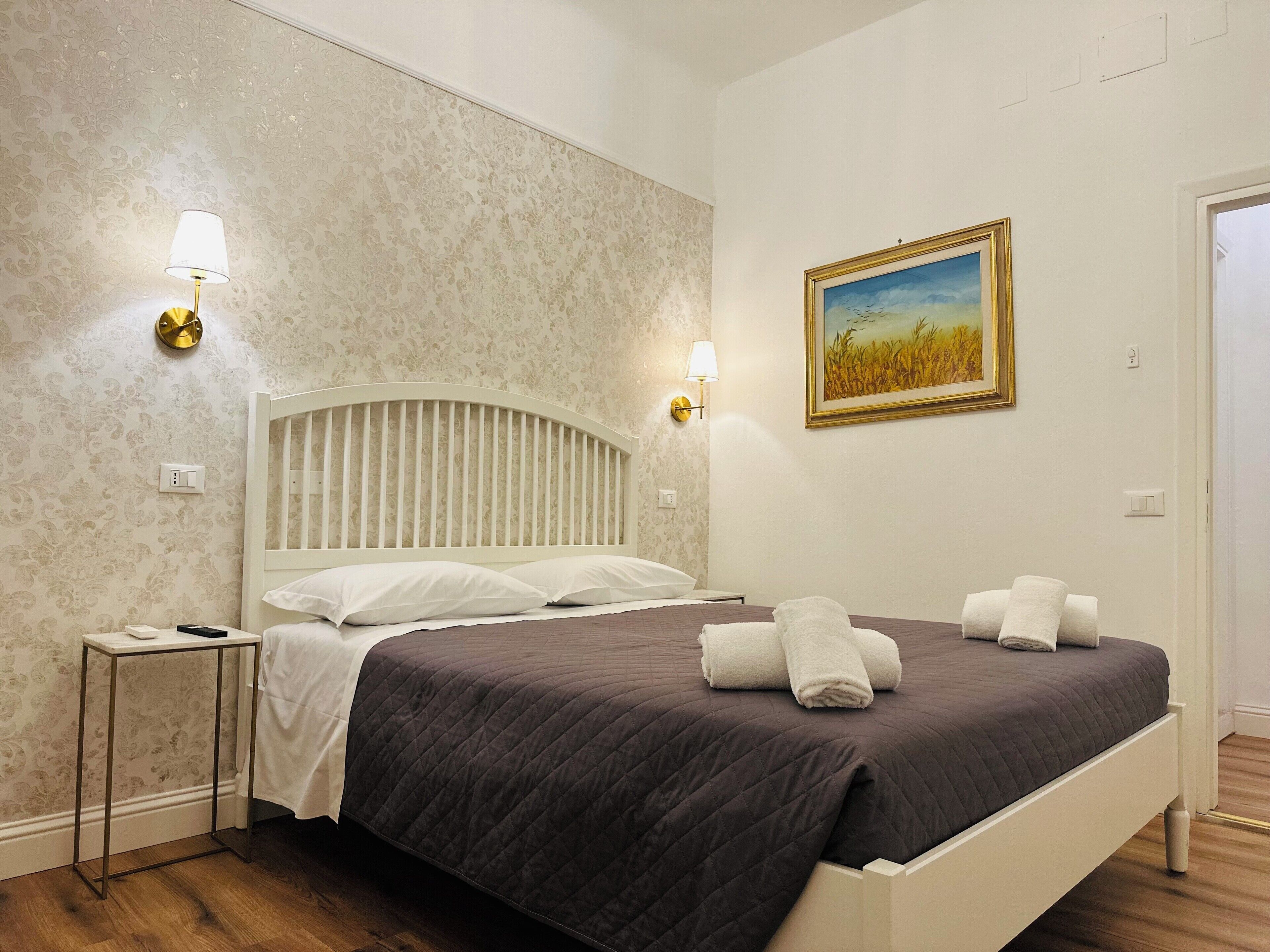 Una cama bien hecha con un edredón oscuro, almohadas blancas y toallas enrolladas sobre un cabecero blanco. Hay una mesita de noche pequeña con una lámpara y un enchufe. En la pared, encima de la cama, se encuentra un cuadro enmarcado de un campo.