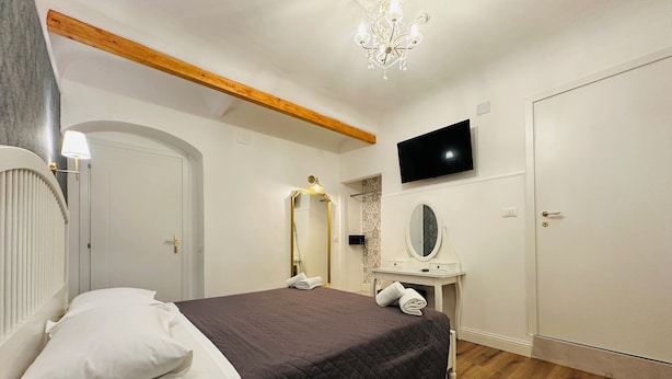 A bedroom with a bed, a vanity with a mirror, a television on the wall, and a wooden ceiling beam. - Relais Sampaoli  (Florence)