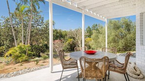 Eine überdachte Terrasse mit einem runden Tisch und Korbstühlen, umgeben von einem Garten mit verschiedenen Pflanzen und Bäumen.