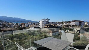 A terrace with a table and chairs, overlooking a coastal town with buildings and mountains in the background. - Adele Hotel Ksamil (Ksamil)