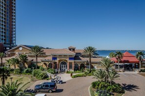Ein Hotel im mediterranen Stil, mit einem zentralen Eingang, der von einer roten Markise, Palmen und Blick auf das Meer geschmückt ist.