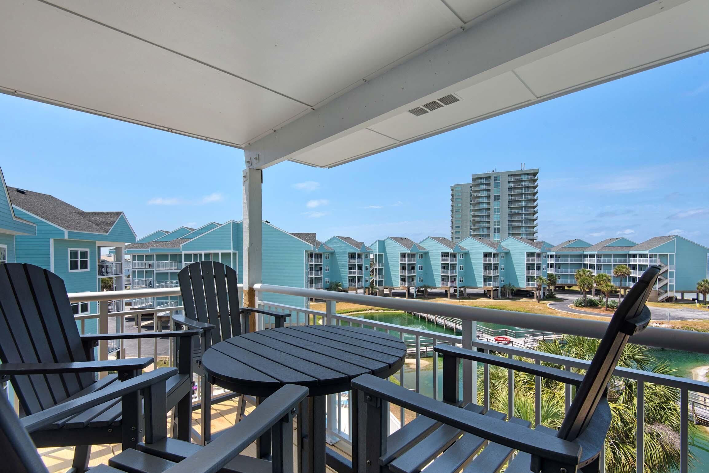 A balcony with Adirondack chairs and a table, overlooking a pool and a row of light blue buildings.