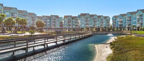 A waterfront hotel with multiple-story buildings, a wooden bridge, and a clear blue pool.