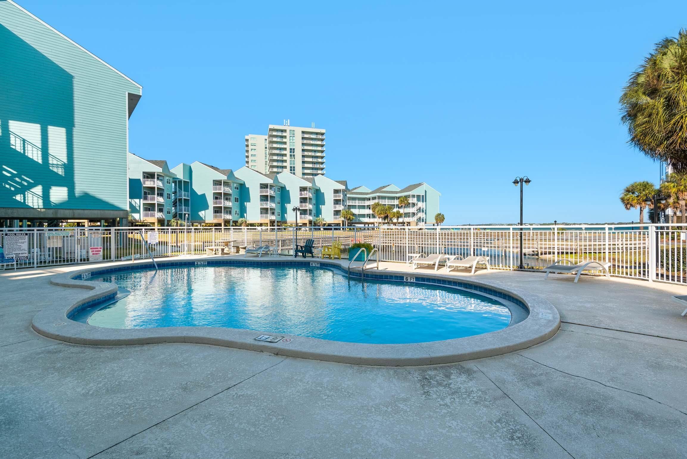 A swimming pool with lounge chairs, a fenced area, and a building with multiple balconies in the background.