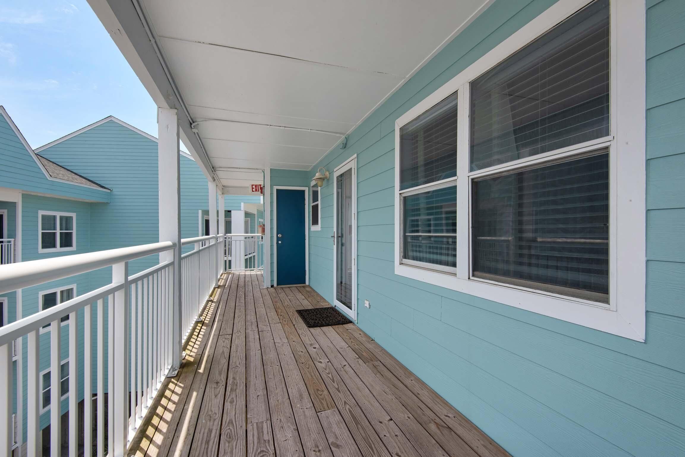 A covered wooden deck with a white railing, a blue door, and a window with blinds.