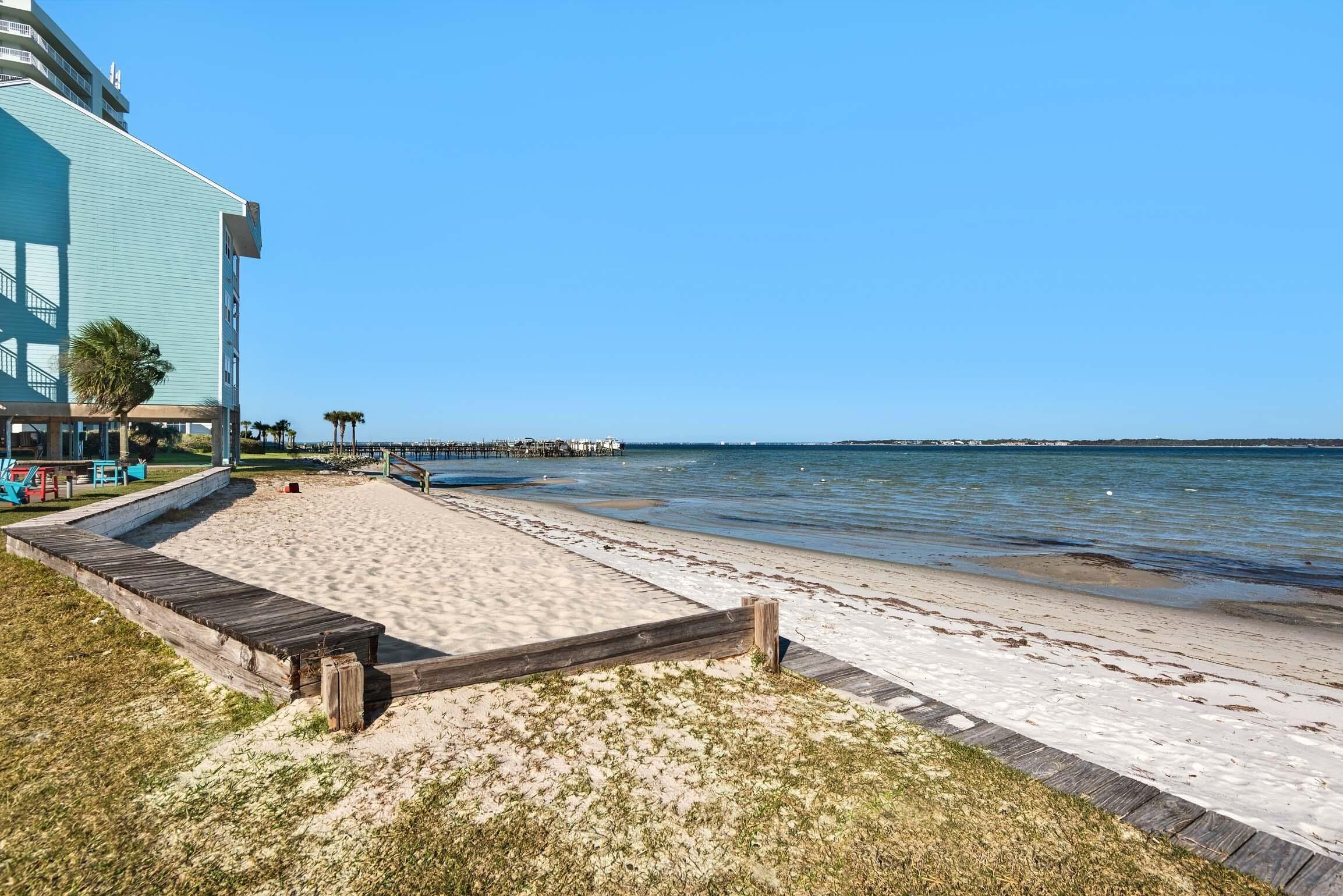 Une plage avec une promenade en bois menant à l’eau, un bâtiment à la façade bleue et une jetée en arrière-plan.