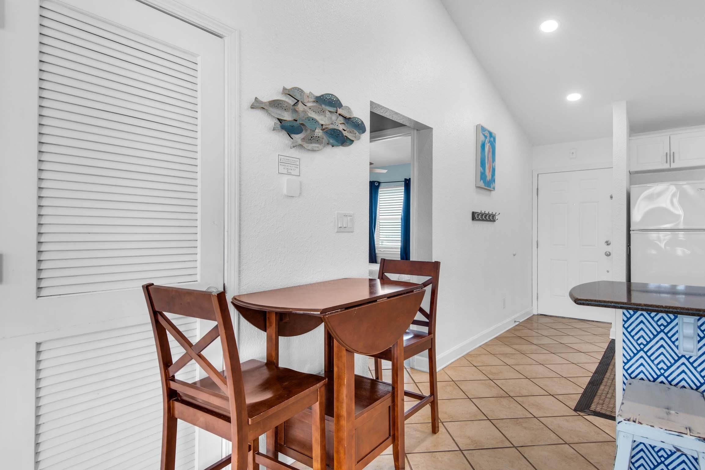 A dining area with a wooden table and chairs, a decorative fish wall art, and a kitchen with white cabinets and a patterned rug.