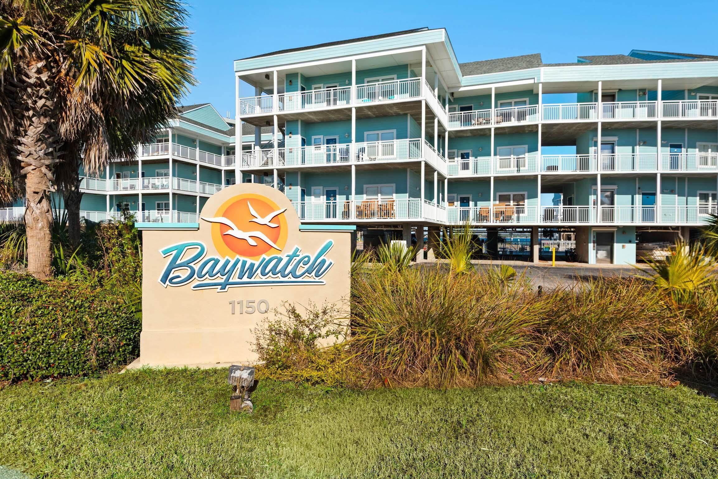 Un hotel de varios pisos con balcones, un cartel que dice "Baywatch 1150" y una palmera en primer plano.