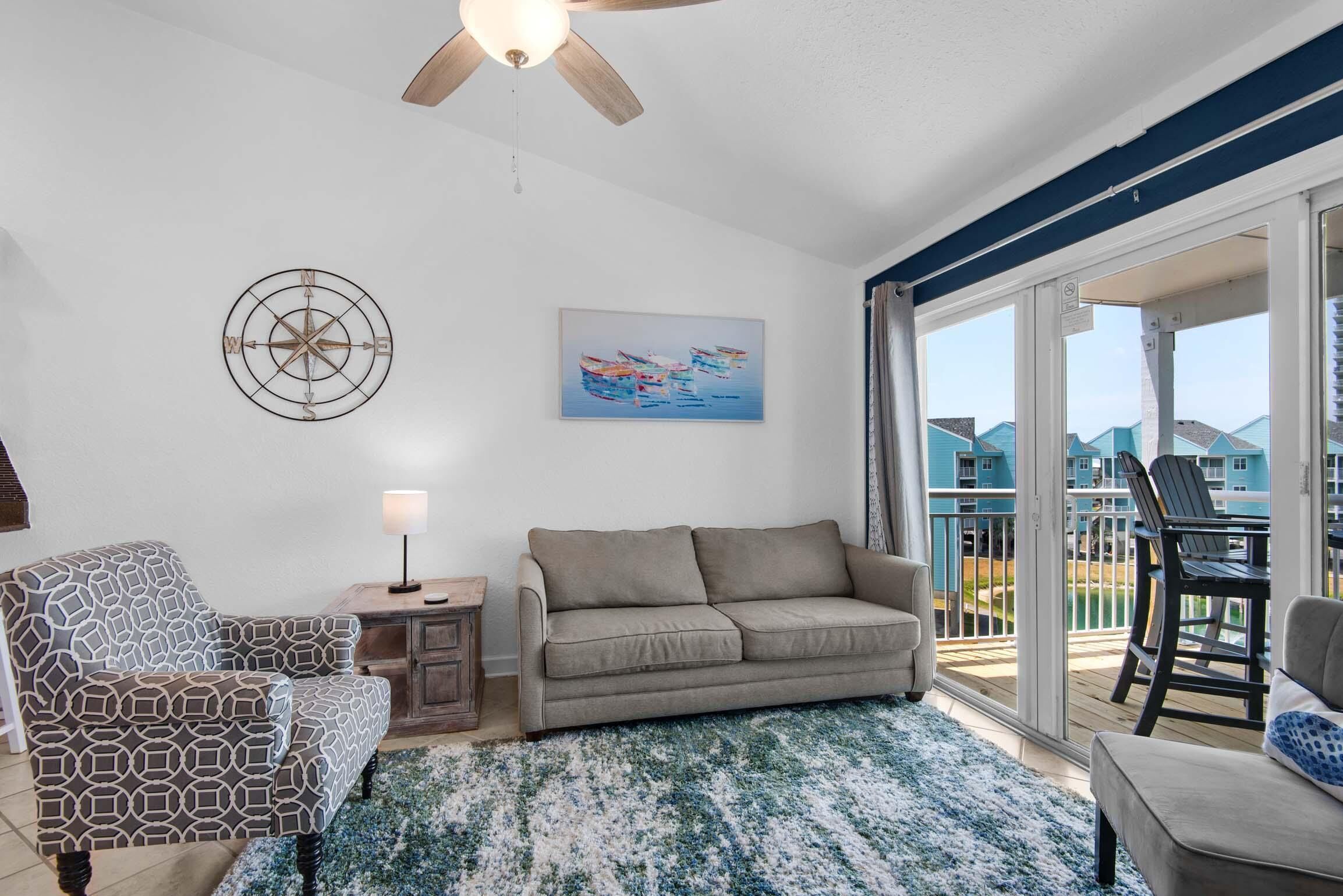 A living room with a sofa, patterned armchair, and a ceiling fan. There is a painting on the wall and a view of a balcony with outdoor furniture.
