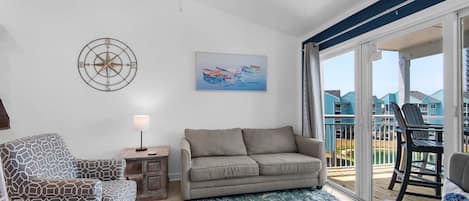 A living room with a sofa, patterned armchair, and a ceiling fan. There is a painting on the wall and a view of a balcony with outdoor furniture.