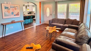 A living room with a sofa, a coffee table, and a guitar. There is a kitchen area with bar stools and a ceiling fan. - Oasis at Muchy Branch (Rehoboth Beach)