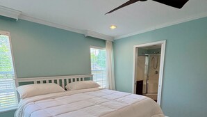A bedroom with a bed, a ceiling fan, a window with blinds, and a door leading to a bathroom. - Oasis at Muchy Branch (Rehoboth Beach)