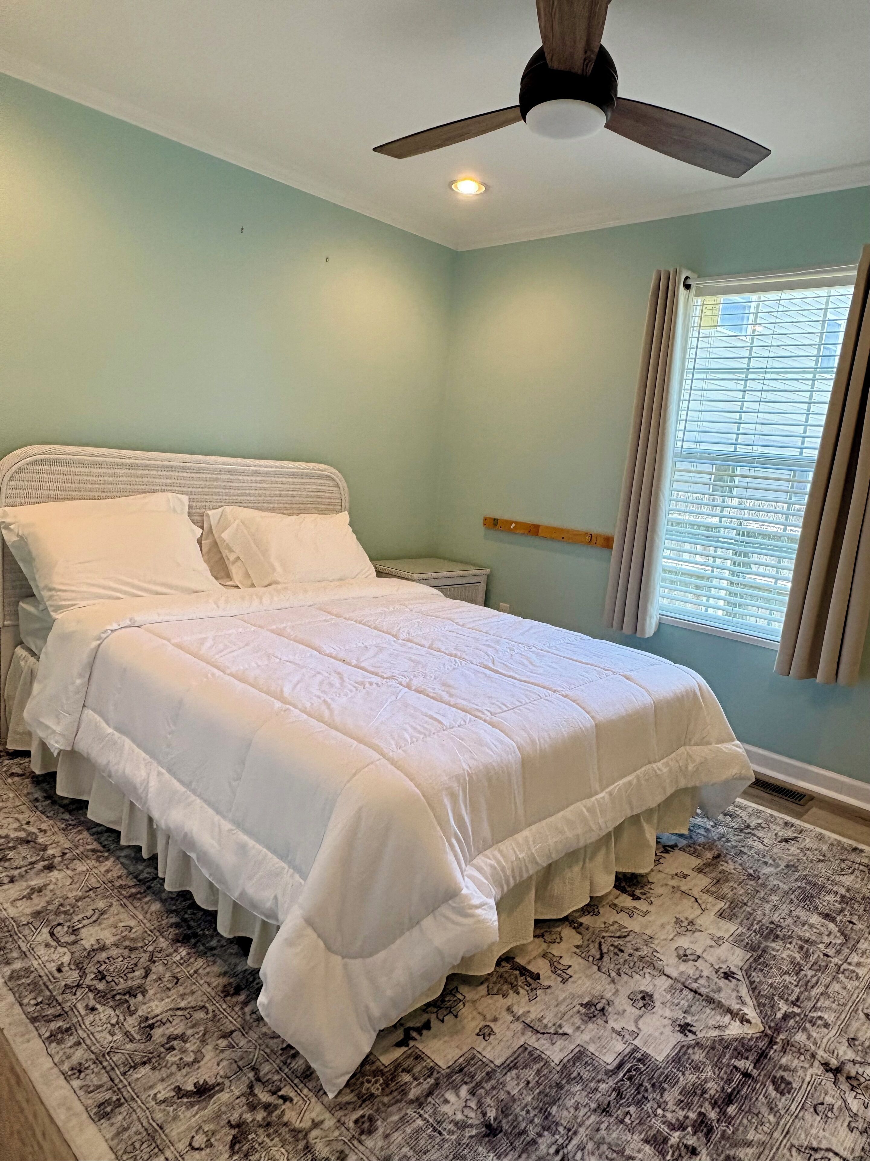 A neatly made bed with a white comforter and pillows, a ceiling fan, a window with blinds, and a patterned rug.