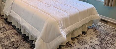 A neatly made bed with a white comforter and pillows, a ceiling fan, a window with blinds, and a patterned rug.