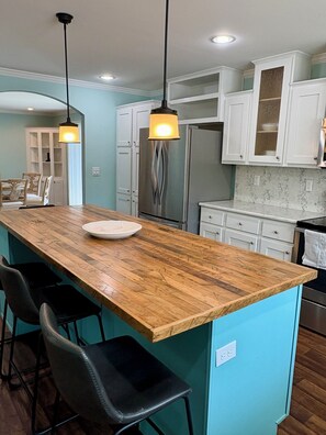 A kitchen with a wooden island, bar stools, white cabinets, and stainless steel appliances. - Oasis at Muchy Branch (Rehoboth Beach)