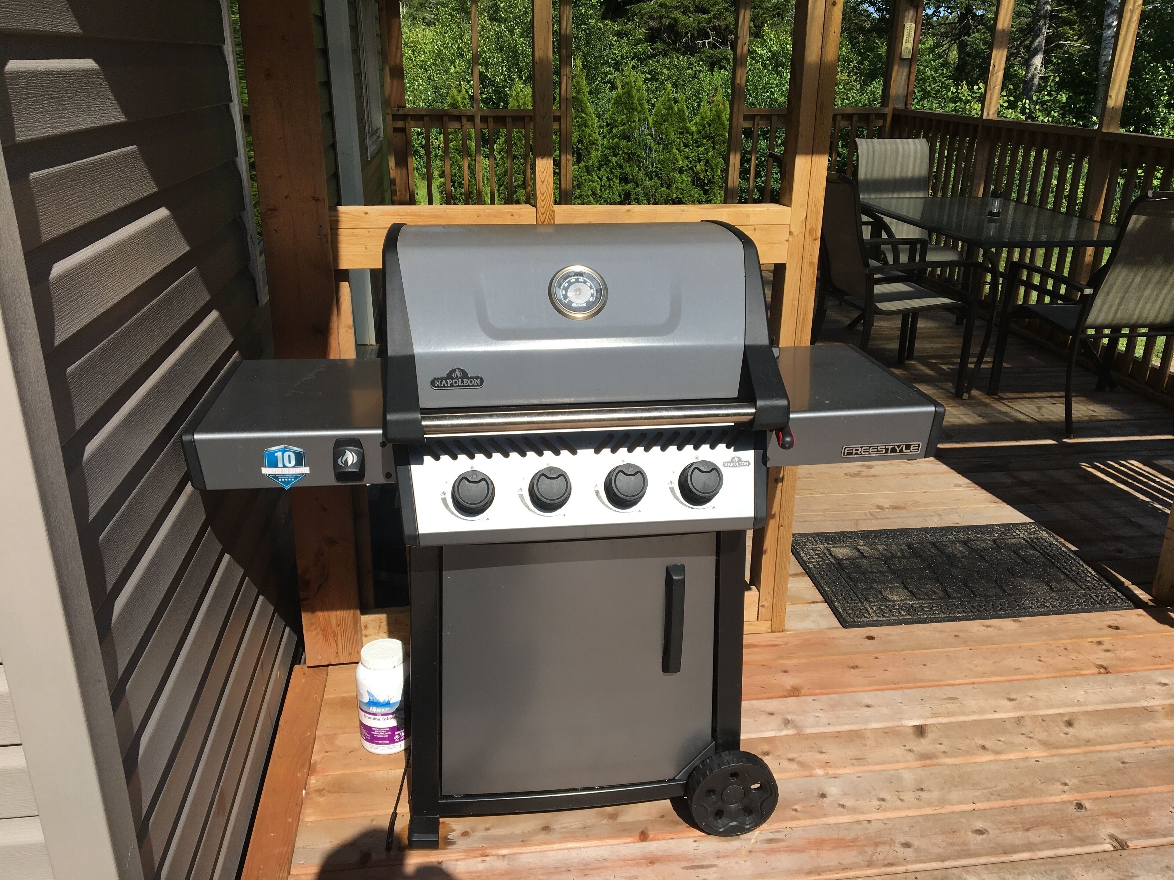 A portable gas grill with controls and a digital display, positioned on a wooden deck with outdoor furniture and a wooden fence in the background.