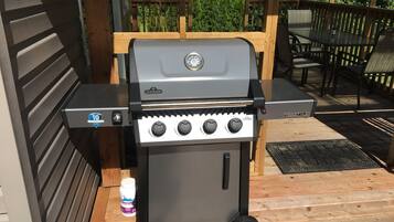 A portable gas grill with controls and a digital display, positioned on a wooden deck with outdoor furniture and a wooden fence in the background.