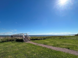 Ein Holzsteg führt zu einer Aussichtsplattform über ein grasbewachsenes Feld, mit blauem Himmel und weiter Wasserfläche im Hintergrund.