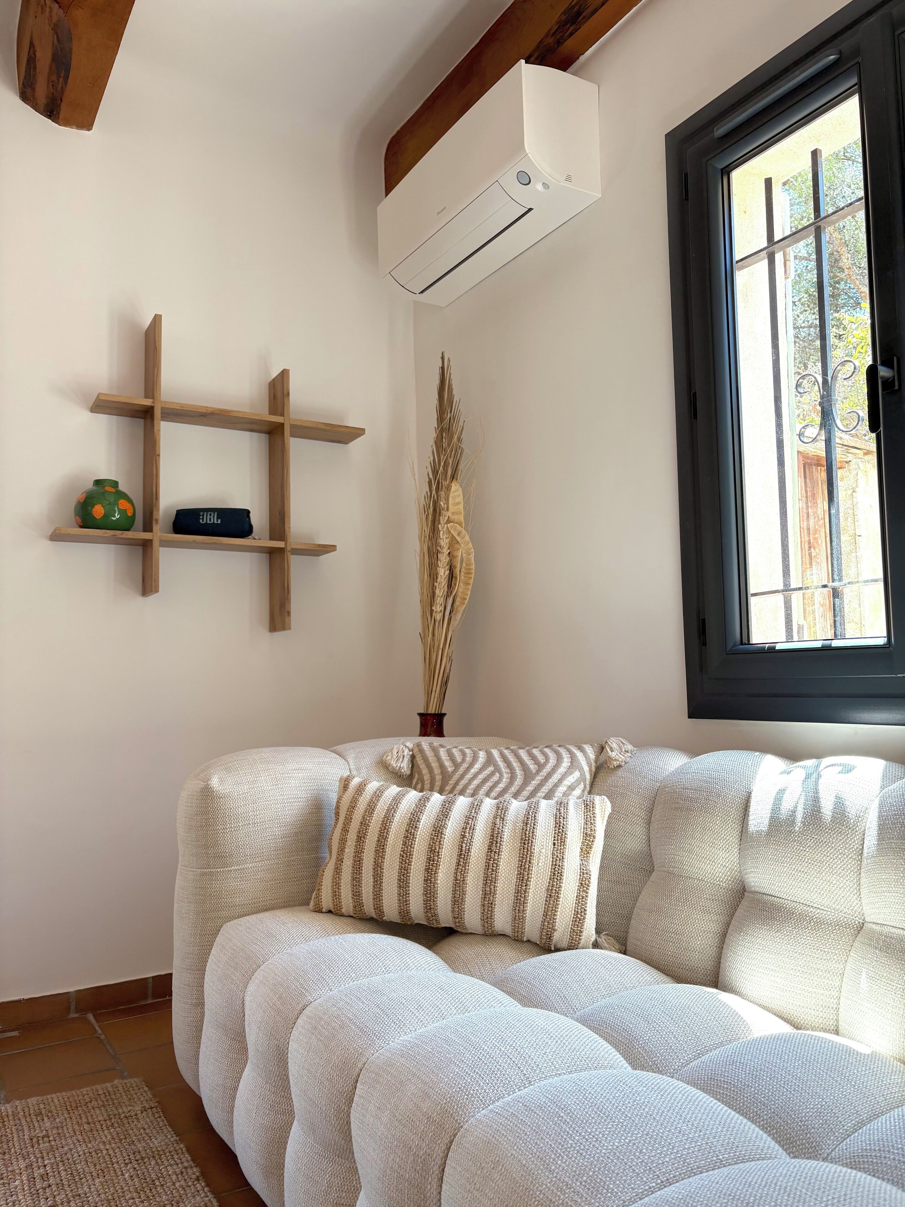 Un salon comprenant un canapé d’angle, une étagère en bois ornée d’objets décoratifs, un appareil de climatisation et une fenêtre offrant une vue agréable.