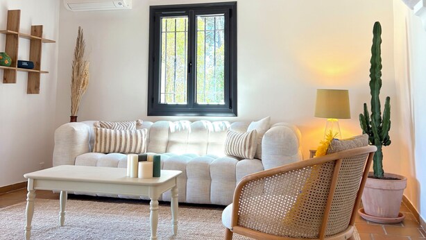 A cozy living room with a sofa, a coffee table, a wicker chair, a cactus plant, and a window with a view of trees. - Beautiful detached villa on one level with pool, view and charging station! (La Cadière-d'Azur)