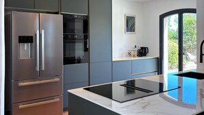 A modern kitchen with a stainless steel refrigerator, built-in oven, and a central island with a black induction cooktop. - Beautiful detached villa on one level with pool, view and charging station! (La Cadière-d'Azur)