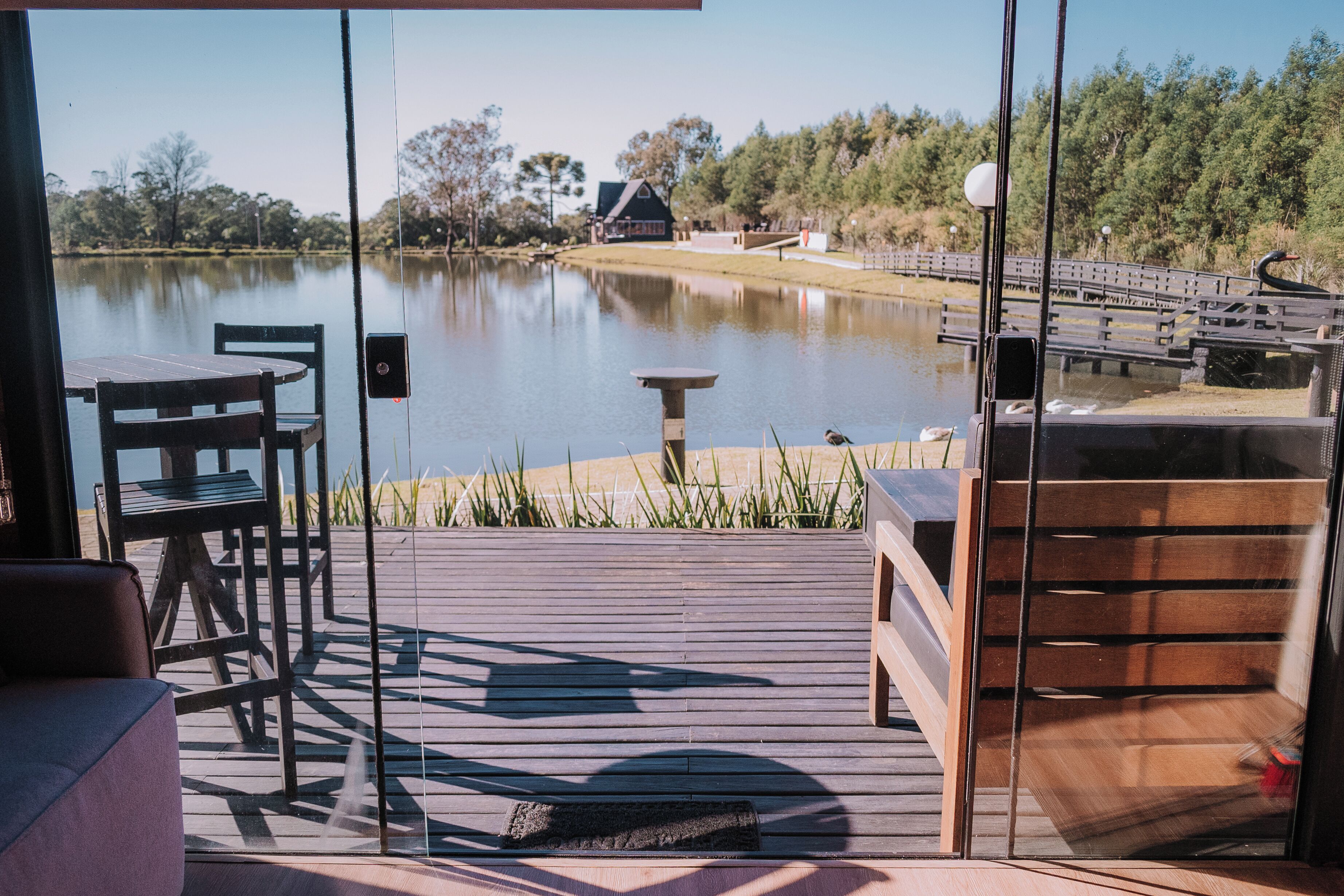 Terraza de madera con una mesa y sillas, con vista a un lago tranquilo con un muelle de madera y una casa al fondo.
